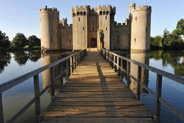 A medieval stone castle with round towers surrounded by a moat, seen from a wooden footbridge across the water